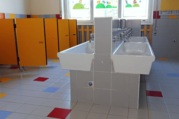 Ein Unisex-Klo in einer Schule, ausgestattet mit hellen, neutralen Farben und modernen Sanitäreinrichtungen. Die Türen der einzelnen Kabinen reichen von Boden bis Decke, um maximale Privatsphäre zu gewährleisten. Außerhalb der Kabinen befindet sich ein gemeinsamer Waschbereich mit Spiegeln und Waschbecken, die für Rollstuhlfahrer zugänglich sind. Über den Waschbecken hängt ein Schild, das an die Wichtigkeit des Händewaschens erinnert, und ein weiteres, das die Unisex-Toilette als einen Raum kennzeichnet, der von jedem unabhängig von der Geschlechtsidentität genutzt werden kann.